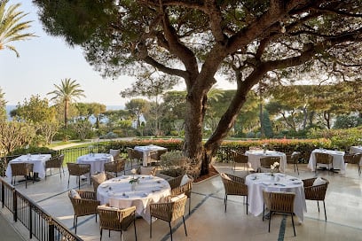 Outdoor dining area at Le Cap restaurant, featuring white tablecloth-covered tables, chairs, lush landscaped gardens, trees and a distant view of the sea