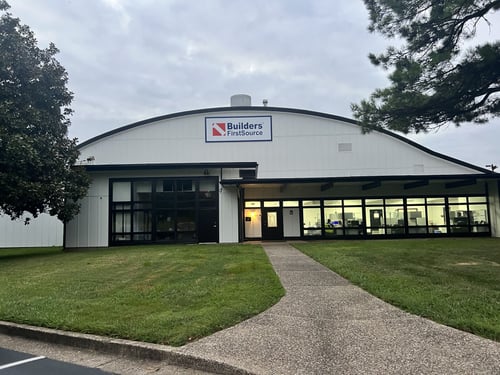 Front view of a white commercial building with a curved roof, large windows, and a sign reading “Builders FirstSource” above the main entrance. A concrete pathway leads across a grassy lawn to the entrance, with trees framing the building on both sides.