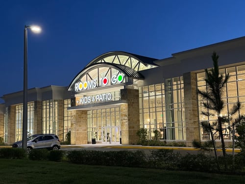 A nighttime view of the Rooms To Go Kids & Patio showroom entrance with modern glass architecture, a glowing sign, and a parked car in the foreground.