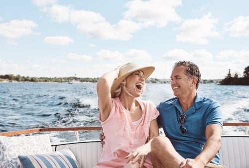 A cheerful couple enjoys a sunny day on a boat. The woman in a pink shirt and straw hat laughs, while the man in a blue shirt smiles back at her.
