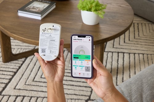 A person holds a smart carbon monoxide alarm next to their phone showing the Advanced Home Health Report on the screen.