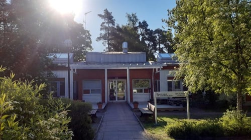 Kuvassa yksikerroksinen, punasävyinen rakennus vehreässä ympäristössä. Kuvassa paistaa aurinko.