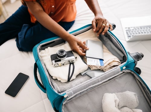 A person packing a blue suitcase with folded clothes, a vintage camera, and travel documents including a boarding pass and passport. Laptop and smartphone are visible.