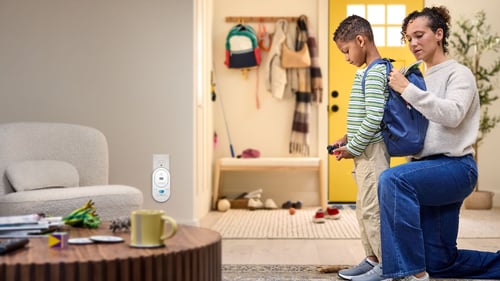 A mom helps her son put his backpack on, and a co alarm is plugged into the background.
