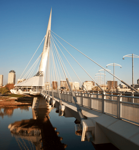 Modern suspension bridge with a tall, sleek spire and cables stretches over a calm river. The city skyline is visible against a clear blue sky.