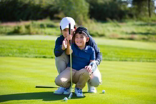 A person in a white cap helps a smiling child line up a golf putt on a sunny course. They are focused, creating a joyful, learning moment.
