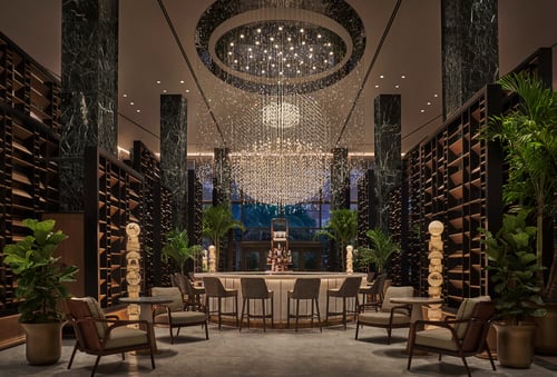 Luxurious hotel bar with a grand chandelier, marble columns, elegant seating, and shelves filled with decor. Large potted plants add a touch of greenery to the space.