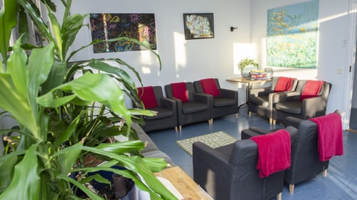 Sisäkuva Breikki Helsingin olohuoneesta. Kuvassa näkyy seitsemän harmaata nojatuolia ja sohva. Nojatuoleilla on pinkkejä vilttejä. Seinillä on tauluja ja takanurkassa on pöytä, jolla on lautapelejä. Huoneessa on myös viherkasveja.