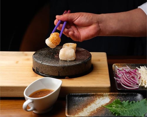 Hand using chopsticks to place a seared scallop on a hot stone, with sauces and garnishes nearby on a wooden board and plates.