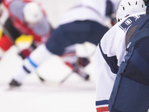 A hockey game scene showing a player in a white jersey in the foreground, with three players clashing in blurred motion behind, conveying dynamic action.