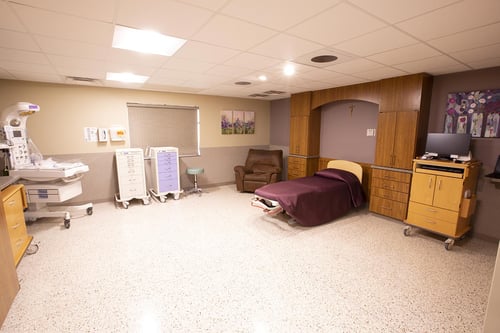 The labor and delivery room with a bed, pull-out sofa, storage cabinets, TV and bassinet.