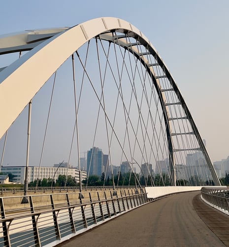 Modern arched bridge with cable suspension spanning over a urban landscape, featuring a cream-colored arch structure, metal cable stays, pedestrian walkway with railings, and a city skyline with tall buildings visible in the background under a clear sky.