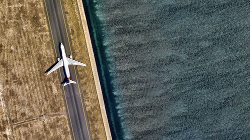 Aerial view of an airplane taxiing on a narrow runway beside shimmering water on the right. Dry, brown grass covers the left side, contrasting the smooth surface.