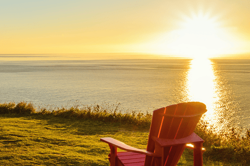 Red Adirondack chair on grassy cliff overlooking peaceful coastal bay at golden sunset.