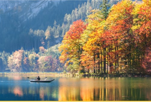 A lone person in a boat glides across a tranquil lake. Vibrant autumn trees reflect in the water, set against a backdrop of forested mountains. Serene and picturesque.