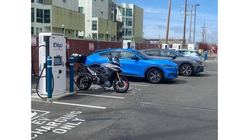 Several EVgo fast charging units installed in marked parking spaces at a public charging station, with EVgo logos on the chargers, protective bollards around each unit. Both NACS and CCS chargers at the station