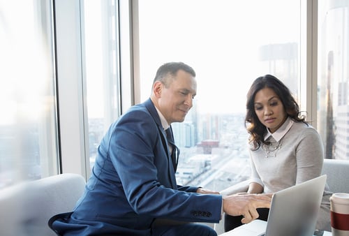 Man in a blue suit and woman in a gray sweater collaborate on a laptop in a modern office with large windows overlooking a cityscape.