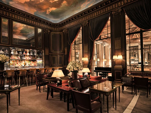 In the center of the photo, we see lamps lit on tables with armchairs and dark red cushions, and a bouquet of white flowers in a vase. On the left is bar 228 with high chairs. We can see the bottles on the shelves. Above the bar is a mirror reflecting the ceiling, which represents a sky with clouds. We look at the woodwork on the walls, with its gilded details. To the right is a piano and large windows with red curtains, revealing another room at Le Meurice, Paris.