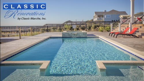 A luxurious backyard pool with clear blue water, surrounded by a wooden deck and two red lounge chairs. Coastal home and dunes in the background.