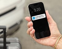 A person holds a phone with a Ring app notification popping up, they look to be outside next to a car with their suitcase.