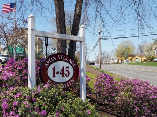 White post sign reading 'Avon Village 1-45 East Main' surrounded by blooming purple azaleas with American flag visible in background, denoting the shopping center that the William Pitt Sotheby's International Realty brokerage is located in Avon, Connecticut.