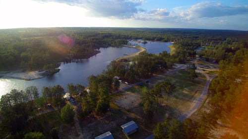 Aerial view of Magnolia Branch Wildlife Reserve with a large lake surrounded by dense forest, several paths and roads, parked vehicles, and small structures. The sun casts a warm glow over the serene landscape.