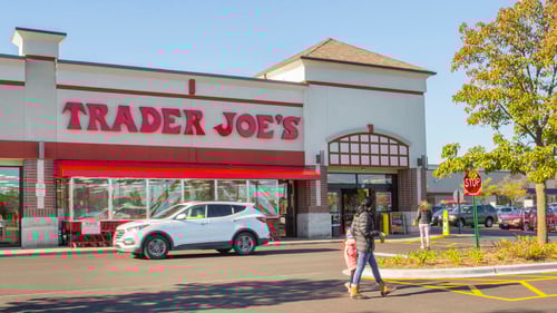 Woman and child in puffy jackets walking past Trader Joe's entrance with a white SUV in front