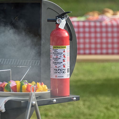 A small fire extinguisher sits on a table extension of a grill, there is smoke rising and a plate of kebabs next to it.