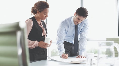 Two professionals collaborate in a modern office, one reviewing documents while the other holds a coffee cup.