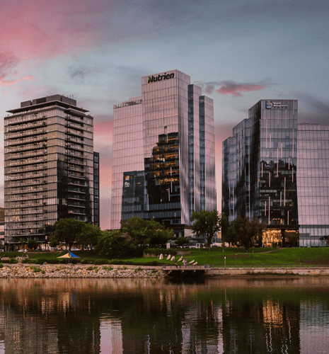 Modern glass skyscrapers reflect a colorful sunset over a calm river, with lush greenery in the foreground, creating a serene urban landscape.