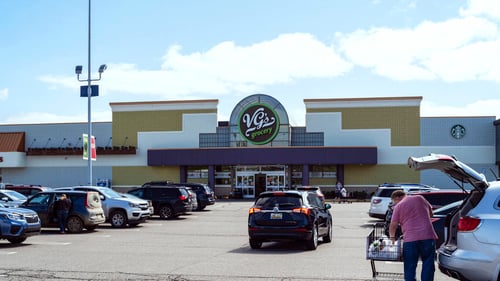 Person in pink shirt putting groceries into back of SUV, black hatchback driving towards front facade of VG's Grocery at Silver Pointe Shopping Center