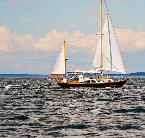 A traditional sailing yacht with dark hull and tall wooden masts, white sails unfurled, sailing on calm blue ocean waters with a small tender alongside, distant shoreline visible under partly cloudy skies.