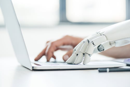 A human and a robotic hand type on a laptop, symbolizing collaboration between humans and technology. The setting is bright, suggesting innovation.