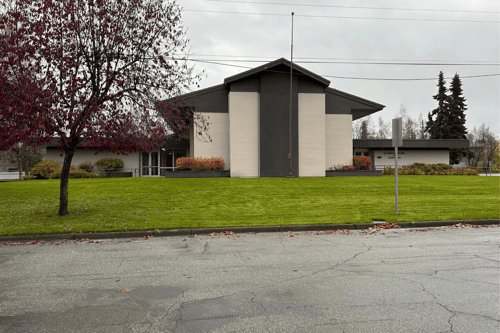 Front view of the Maplewood church building with tall light brick panels and dark trim. A red-leafed tree stands on the left. Both the north and south entrances are visible, with ramps and landscaped shrubs. A wide green lawn extends across the foregroun