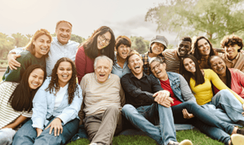 A diverse group of people sitting together on the grass, smiling and posing for a photo in a sunny outdoor setting.