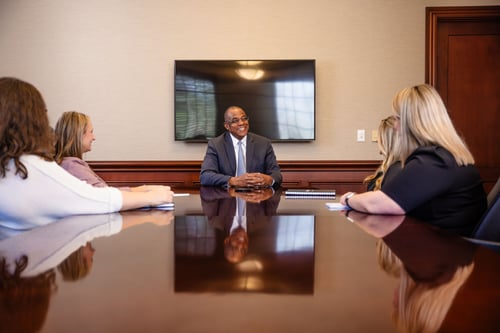 An executive in a suit smiles and leads a discussion with a group of professionals around a large, reflective mahogany conference table. The formal meeting room features warm wood paneling and a large wall-mounted screen.