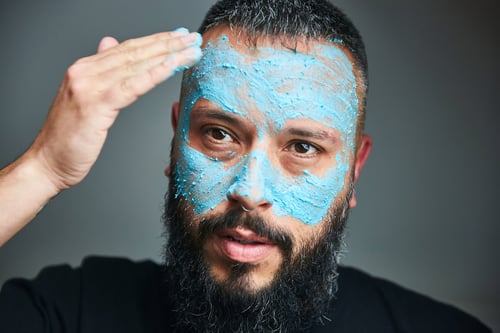 Person with facial hair is applying a blue face mask to their face with their right hand whilst looking at the camera.