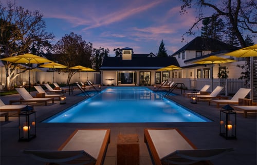 Dusk sky over a rectangular pool lit from within, lined by lounge chairs on either side with a small building at the far end.