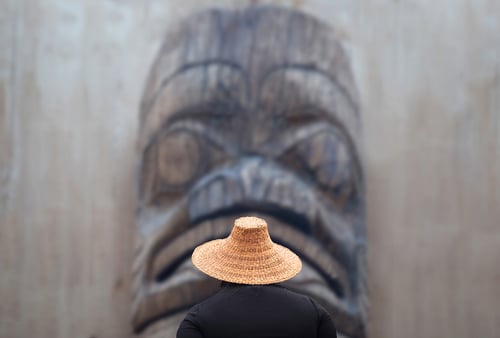 A person in a woven straw hat stands before a large wooden totem pole. The totem's face is detailed, creating a solemn and respectful atmosphere.