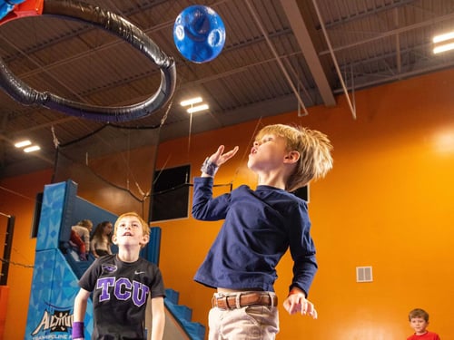 Two boys play indoor basketball, one tossing a blue ball toward a hoop while another watches.