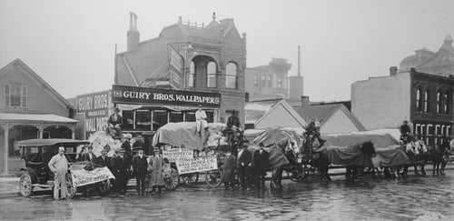 Black and white photo of the original Guiry Bros. Wallpaper Co. store in Denver, with horse-drawn carriages and people gathered outside to see the new 'Spring Stock'.