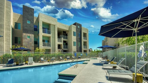 Pool with buildings in the background and patio umbrella up front. Four lounge chairs line the pool.