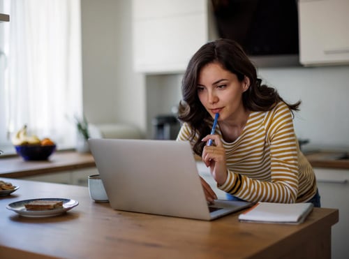 Woman working on laptop at kitchen table while holding pen thoughtfully.