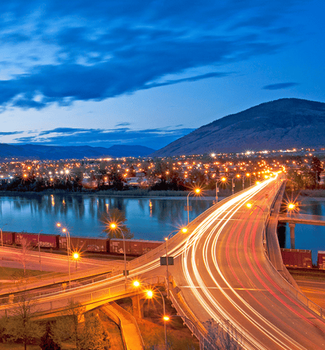 A vibrant night scene shows a bustling, illuminated bridge with light trails from cars. It arches over a river, backed by a distant city and dark hills.