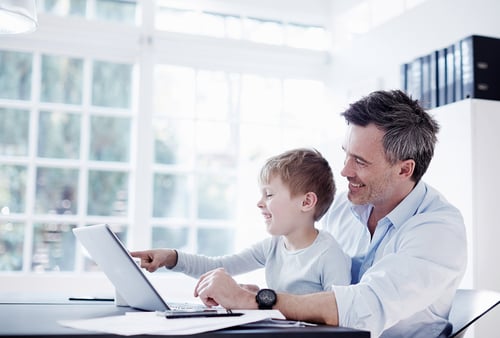 Father and son smiling at a laptop in a bright room with large windows and natural light. The scene conveys happiness and bonding.