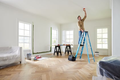 Contractor (man) standing on a ladder installing a dust cover on an alarm on the ceiling. Work table in background.