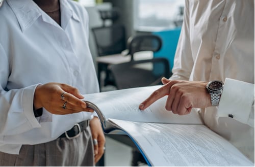 Two professionals in white shirts review a document in an office setting. One points at the page, highlighting details in a collaborative manner.