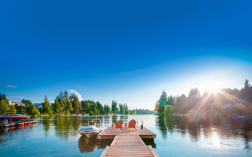 A dock with two red Muskoka chairs at the end, overlooking a lake and surrounding trees on a sunny day.