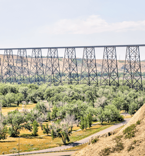 A black steel trestle bridge extends across a lush green tree area, with rolling hills in the background under a clear blue sky. The scene is peaceful and expansive.