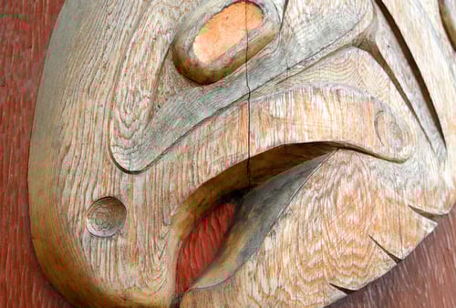 Close-up of a Northwest Coast Indigenous totem pole carving, featuring an intricately carved face with abstract, flowing shapes in natural wood tones.